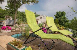 Dos sillones verdes sobre una terraza de madera, con un muro de piedra y una piscina pequeña al fondo.