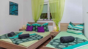 A double bed with green and purple bedding, a bedside table with a lamp, and a window with sheer curtains.