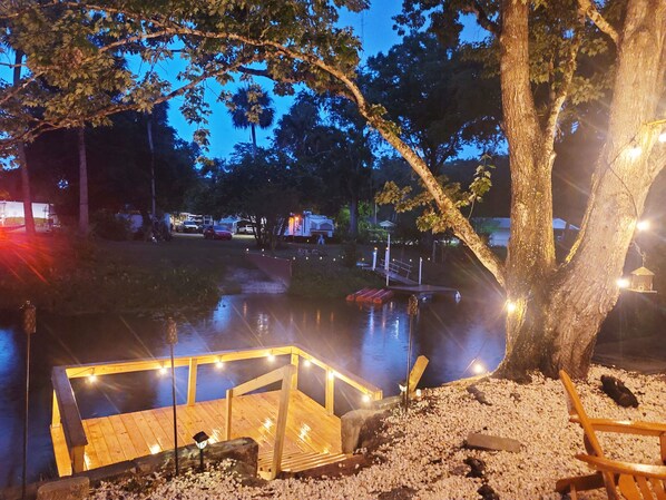 Un espace extérieur aménagé pour la nuit, avec une jetée en bois, un petit bateau et un arbre décoré de guirlandes lumineuses.