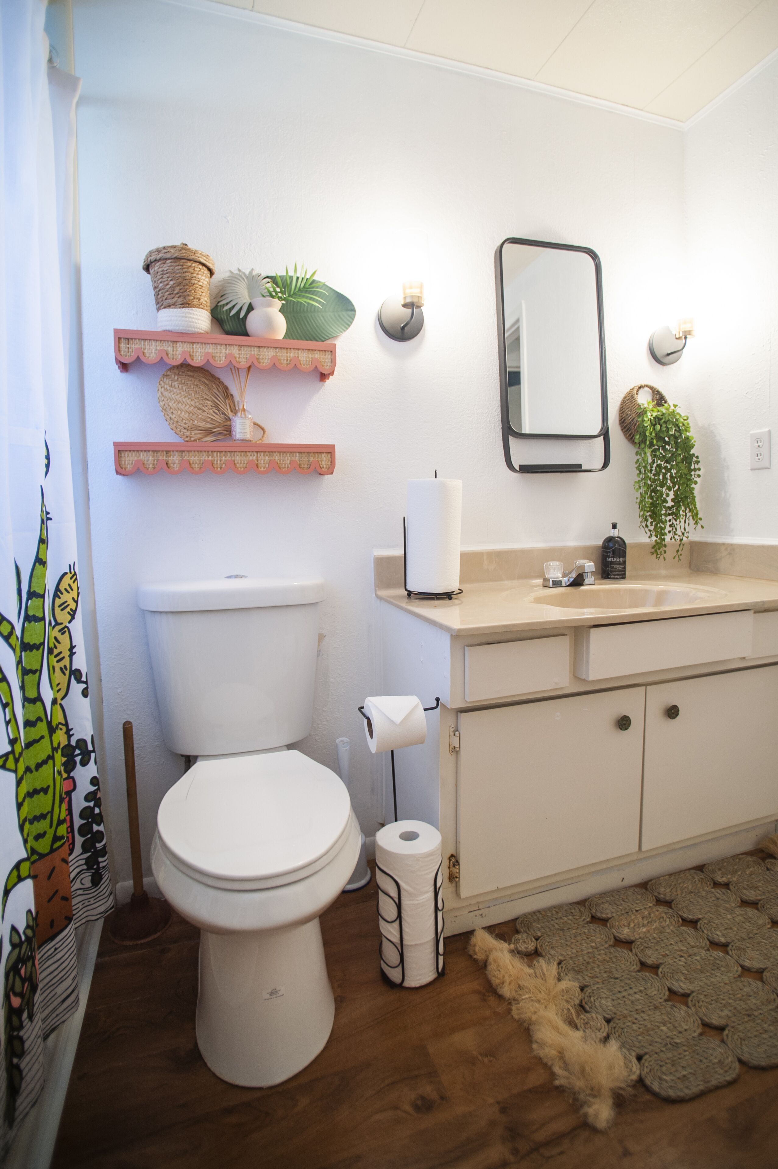Ein Badezimmer mit Toilette, Waschbecken und Spiegel. Auf einem Regal befinden sich Dekorationen, eine Pflanze und ein Duschvorhang.