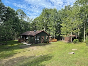 A cabin with a deck, surrounded by a grassy yard and dense forest. - Rickett’s Hidden Treasure- Sullivan County (Lopez)