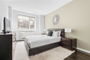 A modern bedroom with a large bed, bedside tables, a TV, and a window with a view of buildings.