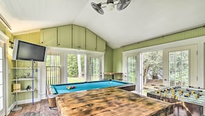 Una mesa de billar, una mesa de fútbolín y una televisión en una habitación con paredes verdes y ventanas amplias.