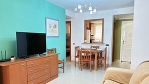 Una sala de estar con un televisor sobre un mueble de madera, un comedor con sillas y una mesa de madera, y una cocina visible a través de un vano abierto.