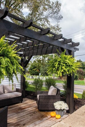 Un espacio de estar al aire libre techado con mobiliario de mimbre y una pérgola, rodeado de vegetación y un cementerio al fondo.