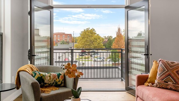 Un soggiorno moderno con un'ampia porta scorrevole in vetro che si apre su un balcone con vista sulla città e sugli alberi autunnali.