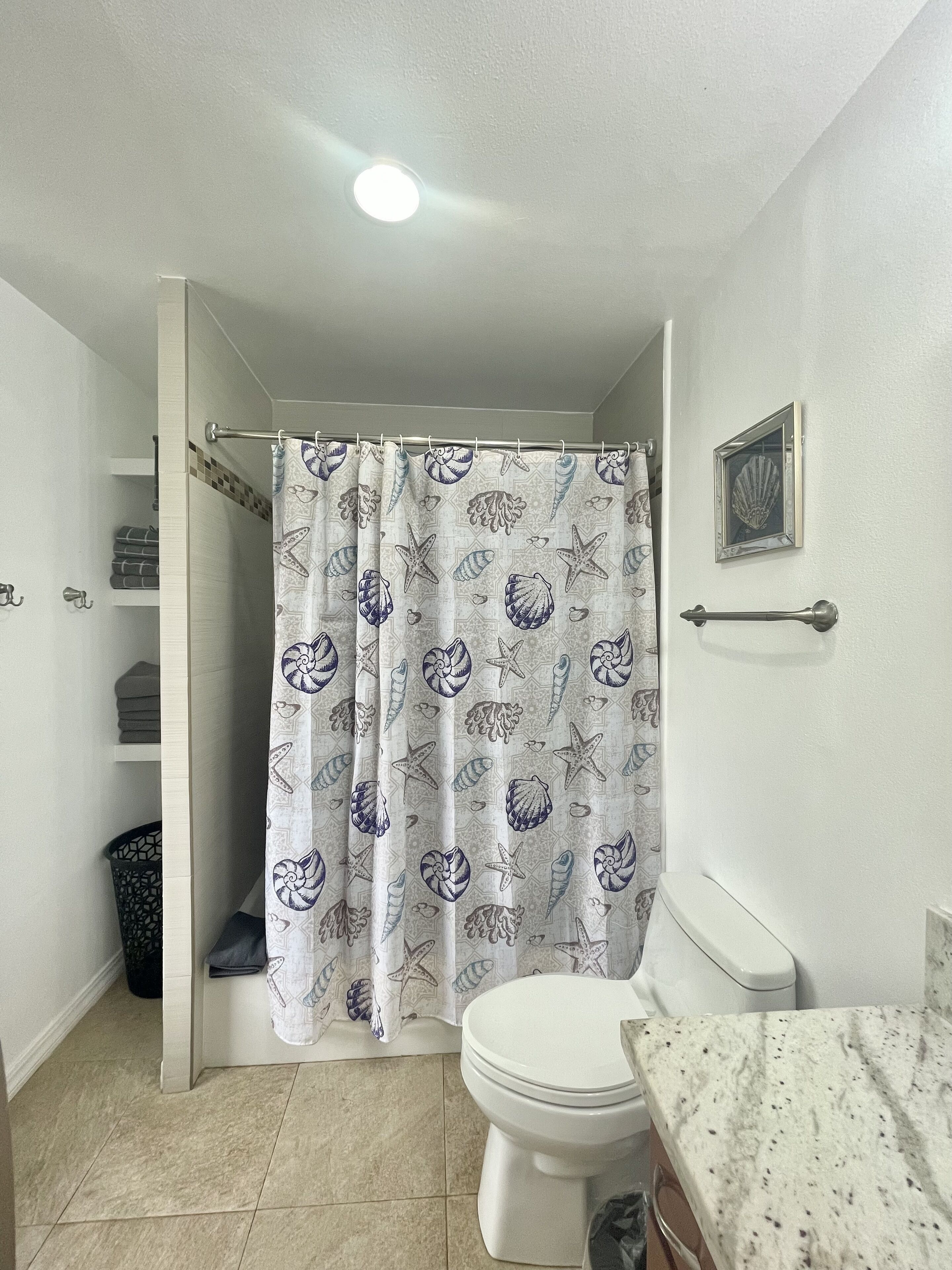 Une salle de bain avec un rideau de douche orné d’un motif représentant des coquillages et des étoiles de mer, des toilettes, un porte-serviettes et un tableau encadré accroché au mur.
