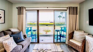 A living room with a sofa, a wicker chair, and a view of the ocean through a sliding glass door. - Beachfront Breeze! Front Row/Heated Pool/Unobstructed Ocean Views!  (Galveston)