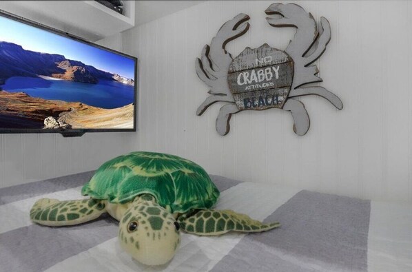 A room with a television displaying a scenic landscape, a decorative wall plaque, and a plush turtle toy on a rug. - Beachfront Breeze! Front Row/Heated Pool/Unobstructed Ocean Views! (Galveston)
