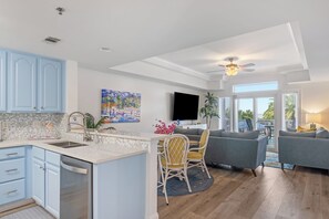 A modern kitchen with a central island, a dining area with a sofa and chairs, a ceiling fan, and a large window with a view of the outdoors.