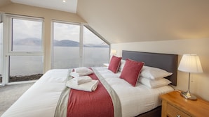 Ein Hotelzimmer mit einem großen Bett, einem Nachttisch aus Holz, einer Lampe und Blick auf das Wasser durch große Fenster.