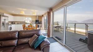 Ein modernes Wohnzimmer mit einer braunen Ledersofa, einem Essbereich und einer Küche. Eine große Schiebeglasdoor führt auf einen Balkon mit Blick aufs Meer.
