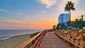 A wooden boardwalk leading to a beach with a palm tree and a tall building in the background. - Calahonda Elegance With Pool And Sea View, Mijas Costa, Spain (Mijas Costa)