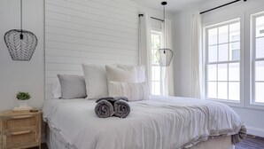 Una cama bien hecha con toallas enrolladas, una mesita de noche con una planta y una ventana con cortinas.
