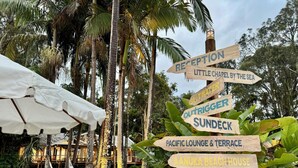 A wooden signpost with multiple directions to various amenities including a reception, little shack by the sea, saltwater outrigger sundock, Pacific lounge & terrace, Aanuka beach house, and surf shack cafe. - Reef @ Aanuka Beach Resort Pool / Spa / Tennis (Coffs Harbour)