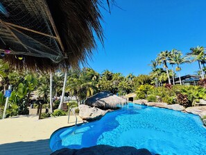 En klarblå pool med en takkonstruktion av halmtak, omgiven av palmer och stenar.