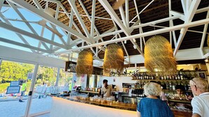 A bar area with a high ceiling and hanging light fixtures, a counter with a variety of bottles, and a view of the outdoors. - Reef @ Aanuka Beach Resort Pool / Spa / Tennis (Coffs Harbour)