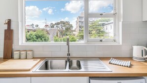 A kitchen with a large window, a stainless steel sink, a wooden cutting board, and a kettle. - Bright & Peaceful Top-Floor Retreat with Harbour Views (Manly)
