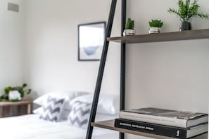 A neatly organized bedroom with a bed, pillows, a bedside table with a plant, and a bookshelf with books.
