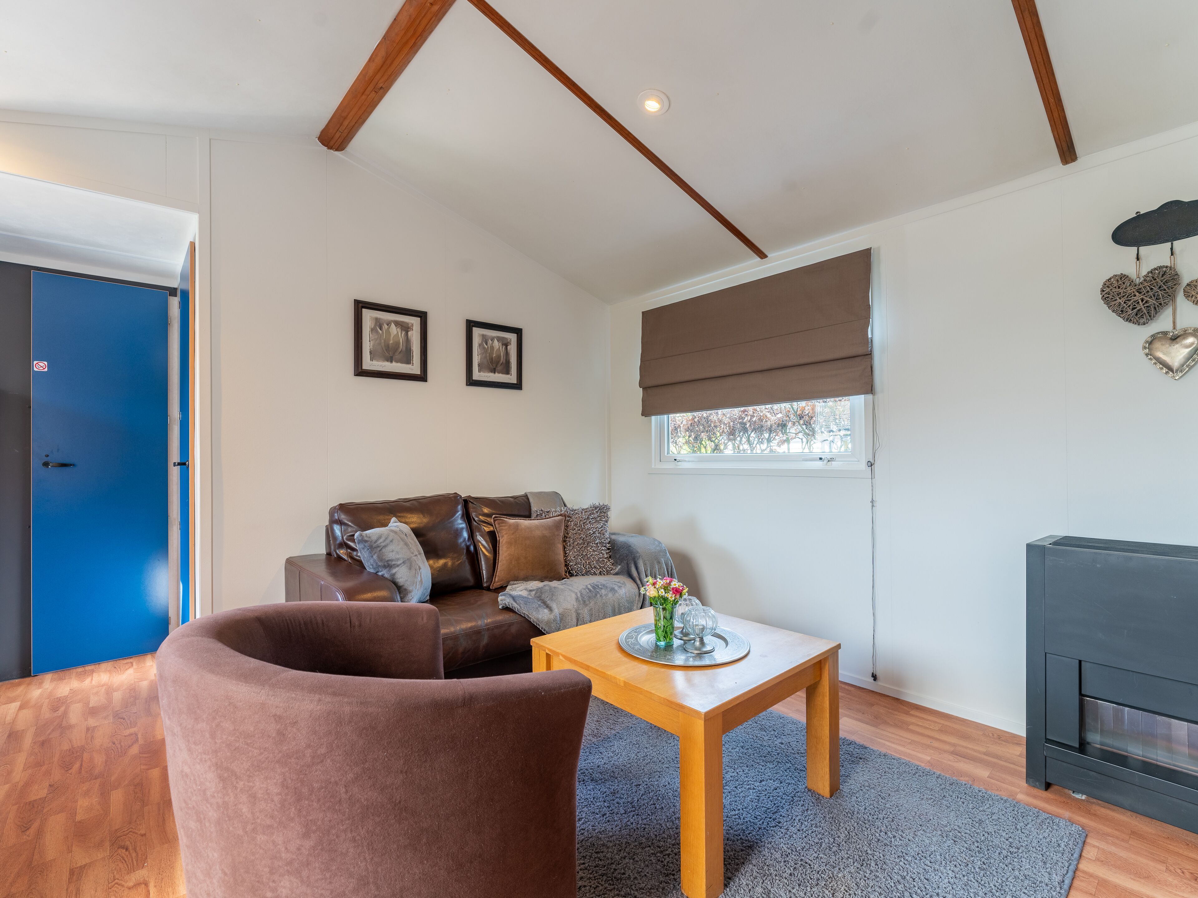 Een gezellige woonkamer met een bruine leren bank, een houten salontafel en een grijs vloerkleed. Er is een blauwe deur, een raam met een bruin zonnescherm en een open haard.