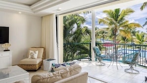 Ein Zimmer mit einer Couch, einem Sessel, einem Fernseher und einem Balkon mit Pool und Palmen.