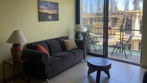 A modern living room with a dark sofa, two side tables with lamps, a painting on the wall, and a large sliding glass door leading to an outdoor area with construction materials.