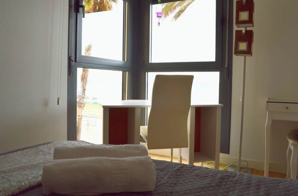 Una camera da letto con un letto, una scrivania e una sedia. Si vede una finestra con vista su palme e un edificio.