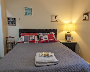 A neatly made bed with a quilt, two pillows, and a folded towel on the bed. There is a nightstand with a lamp and a sign, a chair, and two wall decorations.