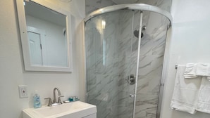 Ein Badezimmer mit einer gläsernen Duschkabine, einem weißen Waschtisch mit Waschbecken und einem Spiegel darüber.
