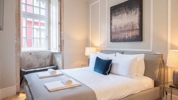 Ein ordentlich bezogenes Bett mit weißen und blauen Kissen, ein Nachttisch mit Lampe und ein großes abstraktes Gemälde an der Wand.