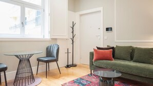 Ein Wohnzimmer mit einem grünen Sofa, einem runden Glastisch, zwei Stühlen und einer Stehlampe.