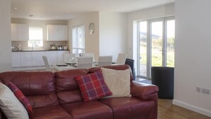 A modern living room with a leather sofa, a dining area with white chairs, and a kitchen in the background.