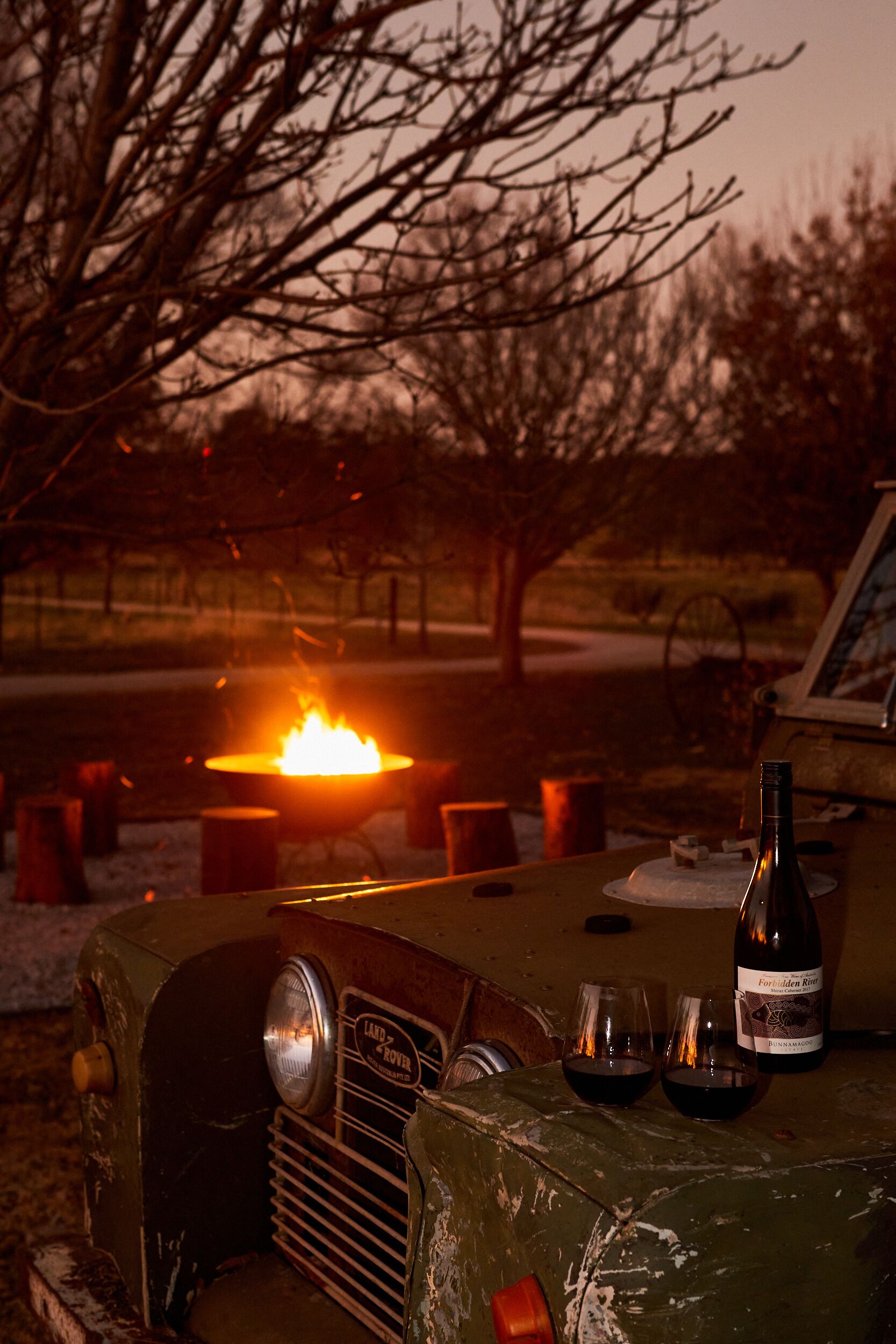 A vintage car with a bottle and glasses on the dashboard, a fire in the background, and a tree.