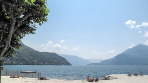 Ein Strand mit freiem Blick auf einen See, umgeben von Bergen. Menschen entspannen sich im Sand, und links ist ein Baum zu sehen.