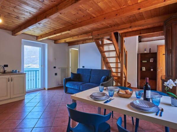 Uma sala de estar ampla com teto de madeira, um sofá azul, uma mesa de jantar posta com pratos e copos, e vista para o mar através de uma grande janela.