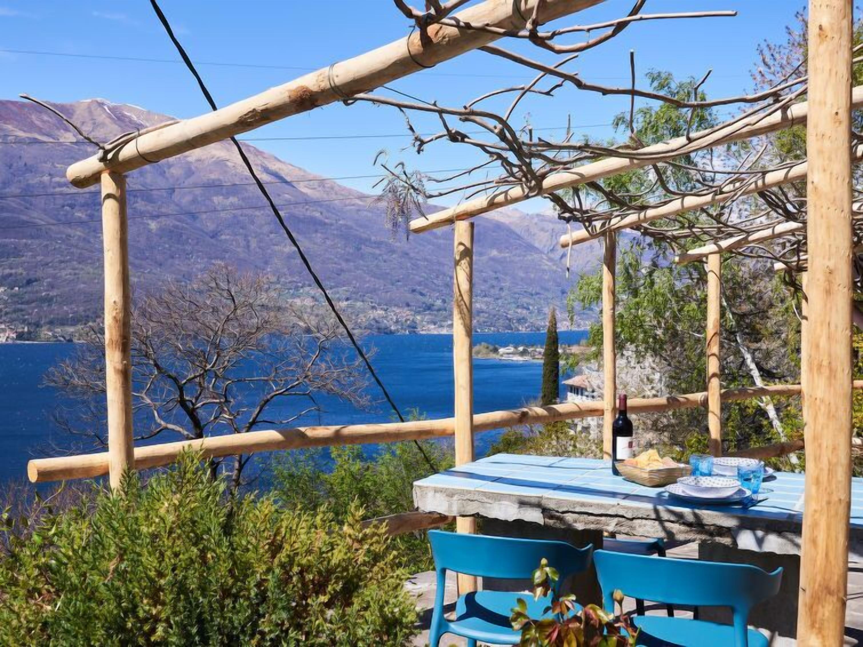 Una pérgola de madera con una mesa y sillas, con vista a un lago y montañas.