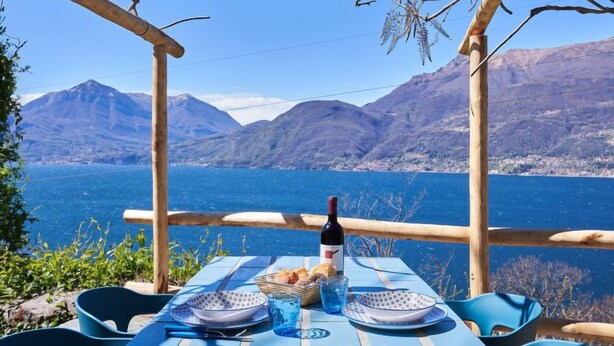 Une table dressée à l’extérieur, avec une bouteille, du pain et des assiettes, donnant sur un lac avec des montagnes en arrière-plan.