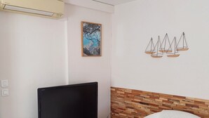 Ein Wohnzimmer mit Flachbildfernseher auf einem Holz-TV-Möbel, einem weißen Sessel mit grünem Kissen, einer wandmontierten Klimaanlage, einem gerahmten Gemälde und einer Wanddekoration mit Segelbooten.