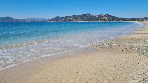 Ein Strand mit klarem, blauen Wasser, Kiesufer und Bergen in der Ferne.