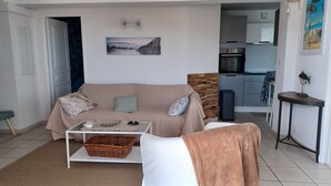 Ein Wohnzimmer mit einem beigefarbenen Sofa, einem Couchtisch und einem Korbwannenmöbel. Im Hintergrund ist eine Küchenzeile mit einer Mikrowelle und einem Spülbecken zu sehen.