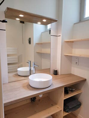 Ein modernes Badezimmer mit einem runden weißen Waschbecken, einem Spiegel und Holzregalen.