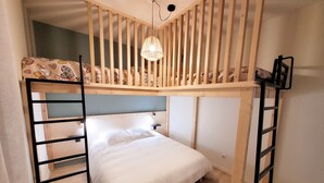 Un lit superposé avec une structure en bois, un matelas blanc et un oreiller à motifs. Il y a une petite étagère et un luminaire suspendu au-dessus du lit.