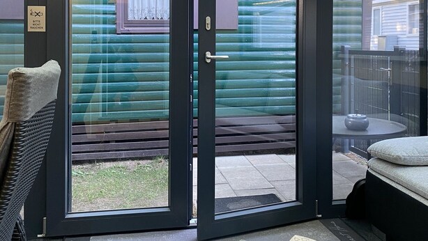 Une porte-fenêtre coulissante donne sur un espace extérieur abrité, doté d’un toit en métal, d’une petite fenêtre avec un rideau et d’un panneau indiquant : « Bitte nicht rauchen » (Merci de ne pas fumer).