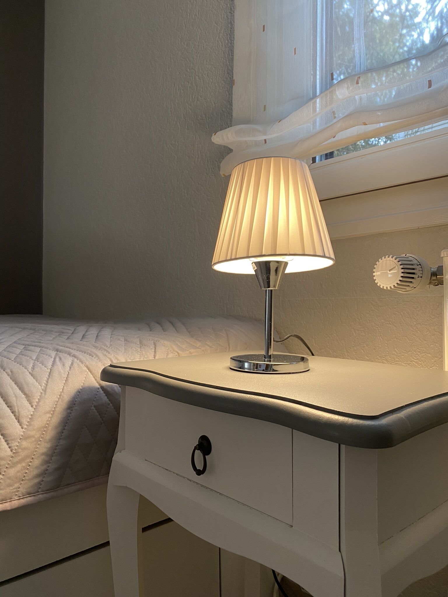 A bedside table with a lamp, a drawer, and a window with blinds.