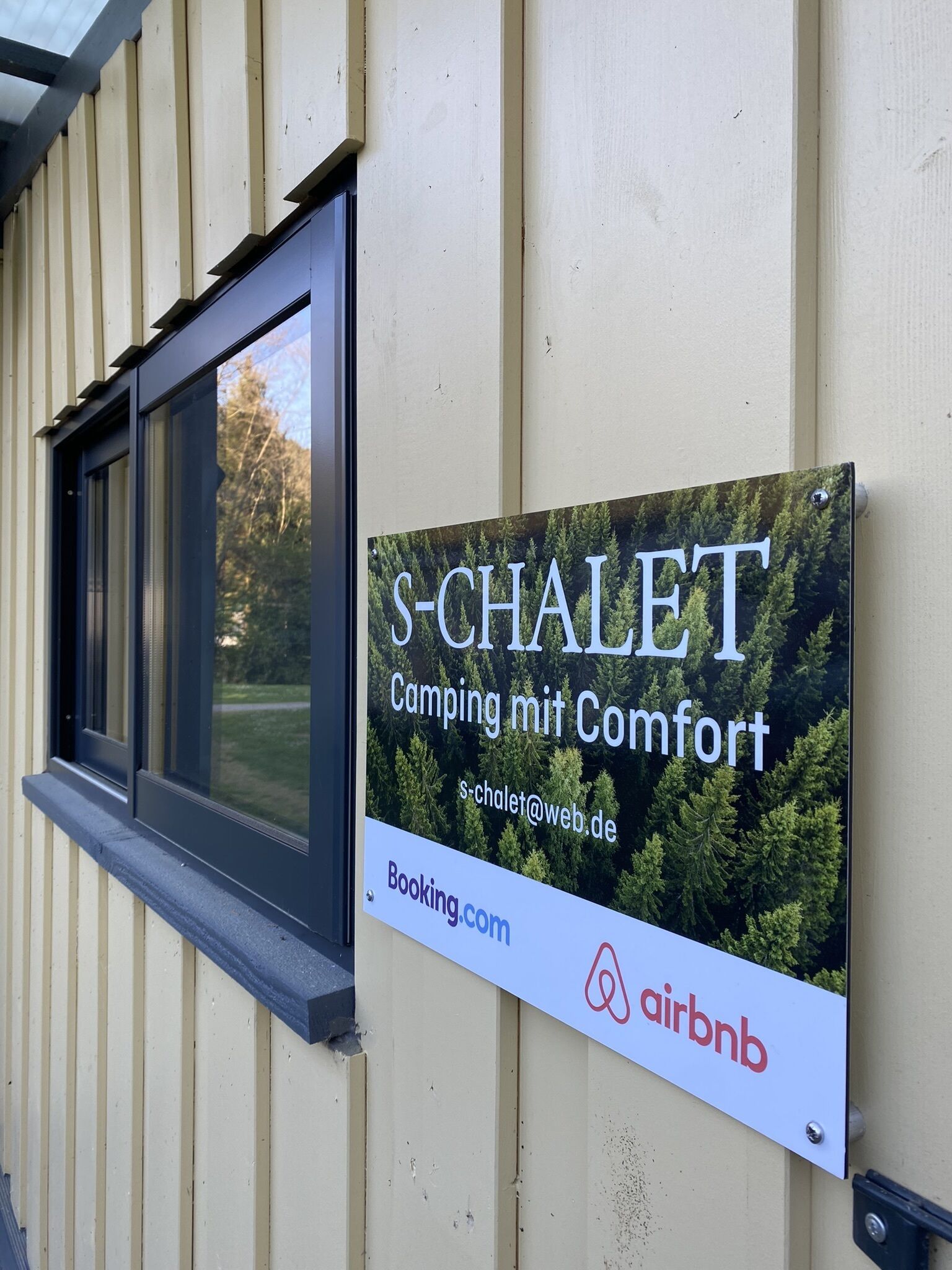 Une enseigne pour le S-Chalet, faisant la publicité d’un camping offrant tout le confort nécessaire, avec un lien vers booking.com et le logo d’Airbnb.