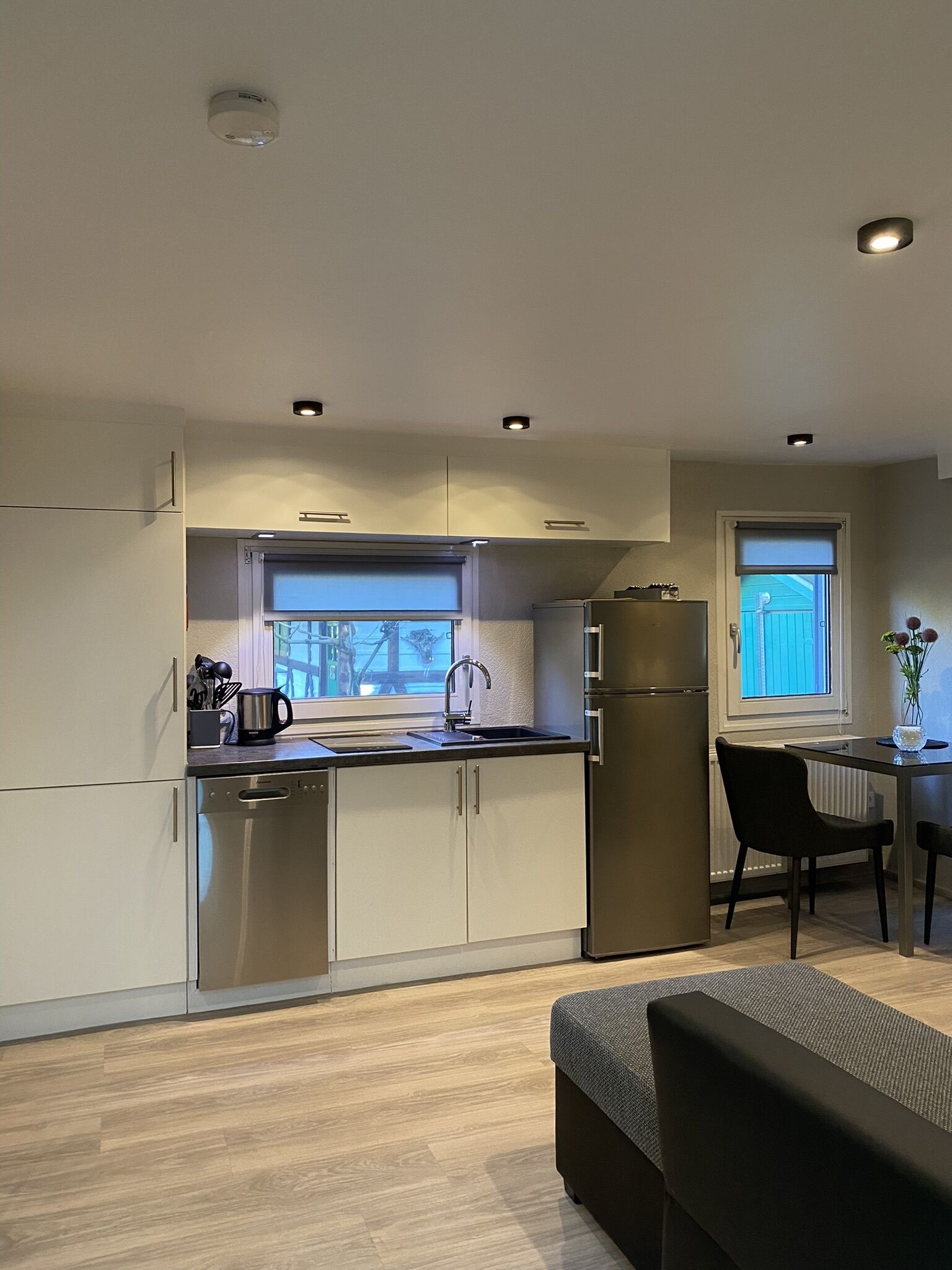 A modern kitchen with a dishwasher, sink, and stainless steel refrigerator. There is a window with a view of the outside, and a dining area with a table and chairs.