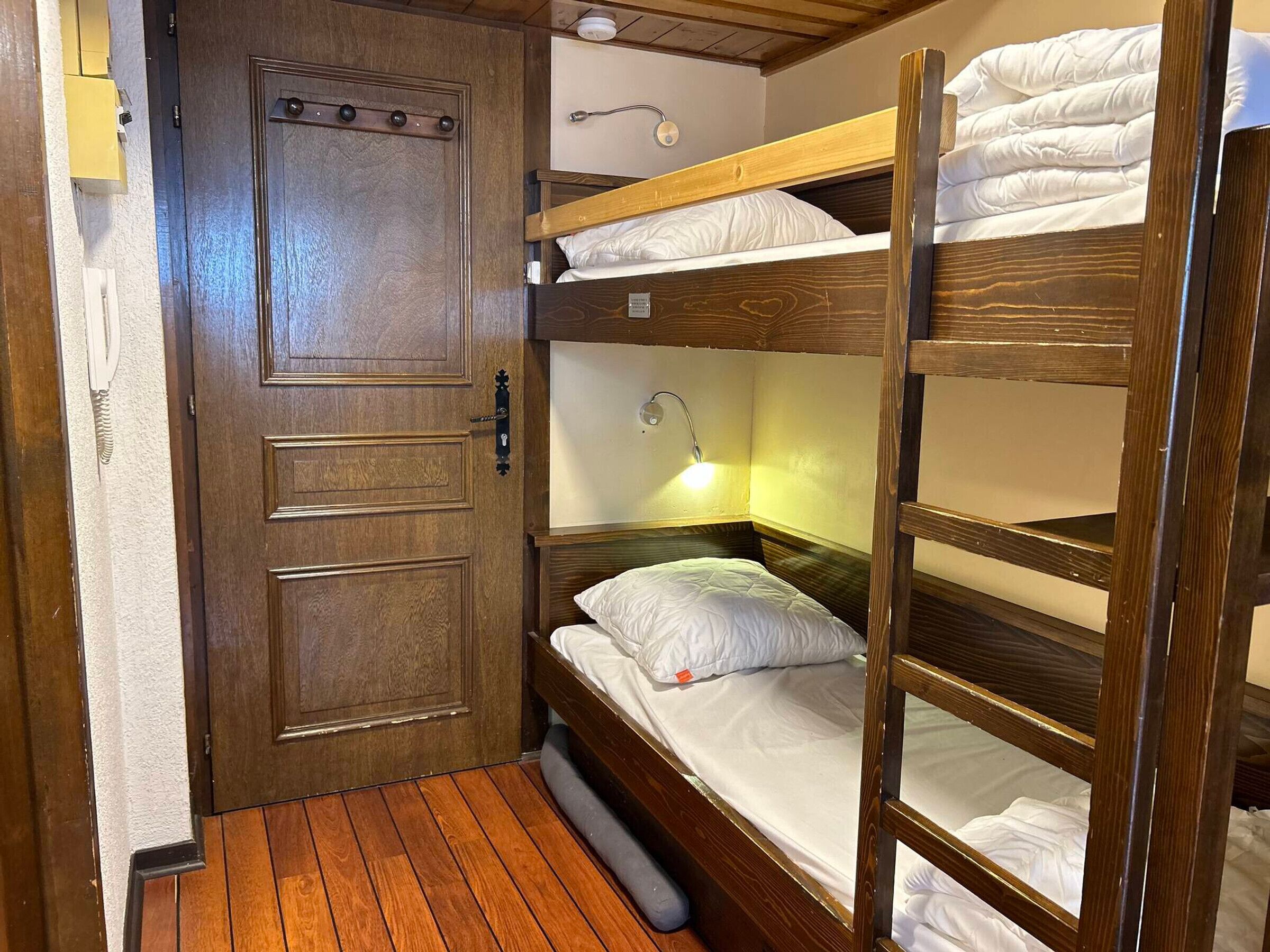 Een kleine kamer met een houten stapelbed, een deur en een nachtkastje.