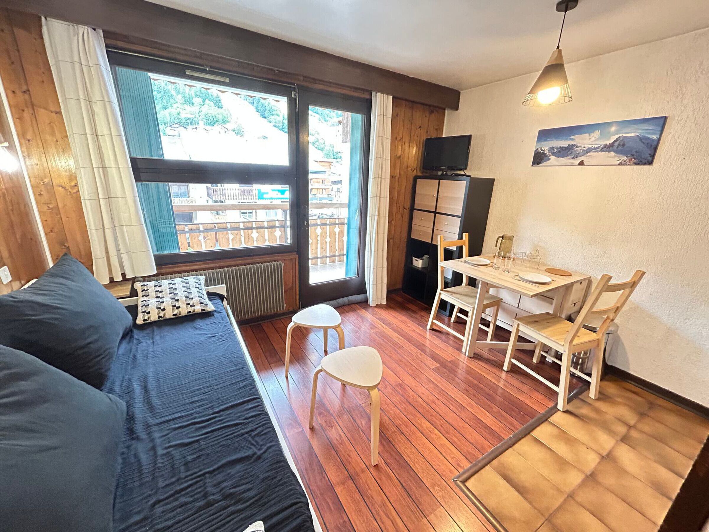 Een gezellige kamer met een houten vloer, een bed met donkere beddengoed, een kleine tafel met stoelen, en een televisie op een kast.