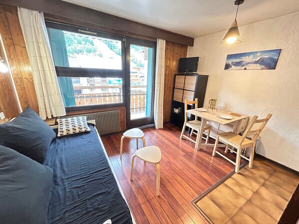 A cozy room with a wooden floor, a bed with dark bedding, a small table with chairs, and a television on a cabinet.