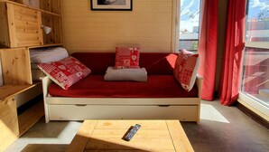 Ein gemütliches Zimmer mit einem Holzbett, einem roten Sofa mit gemusterten Kissen, einem gerahmten Bild an der Wand und einem Fenster mit roten Vorhängen.